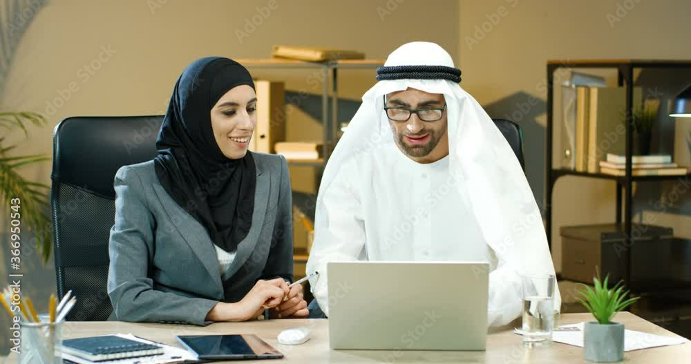 Muslim Arabic businessman and businesswoman working together in office a laptop computer on table. Arab woman in hijab and man boss in dishdasha discussing some project and giving five to each other. Muslim Arabic businessman and businesswoman working together in office a laptop computer on table. Arab woman in hijab and man boss in dishdasha discussing some project and giving five to each other.