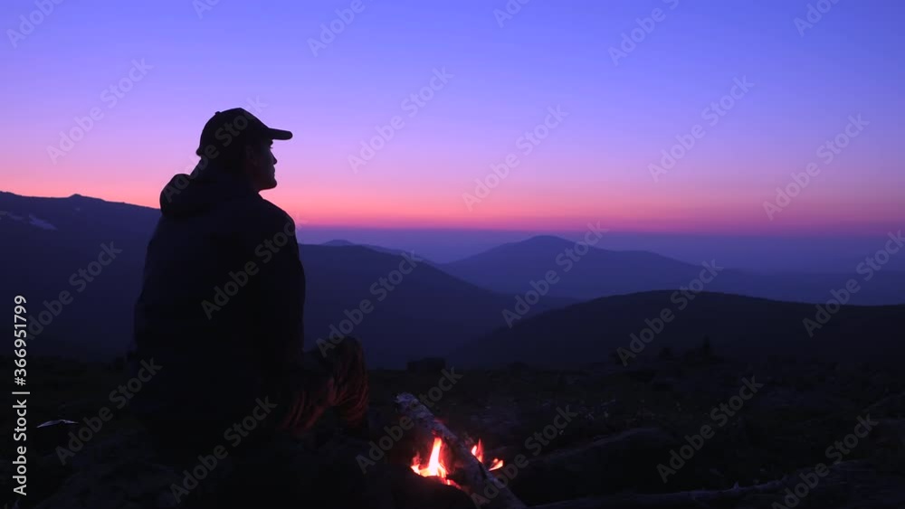 A man sits by a fire in the mountains and looks at the night sky ...