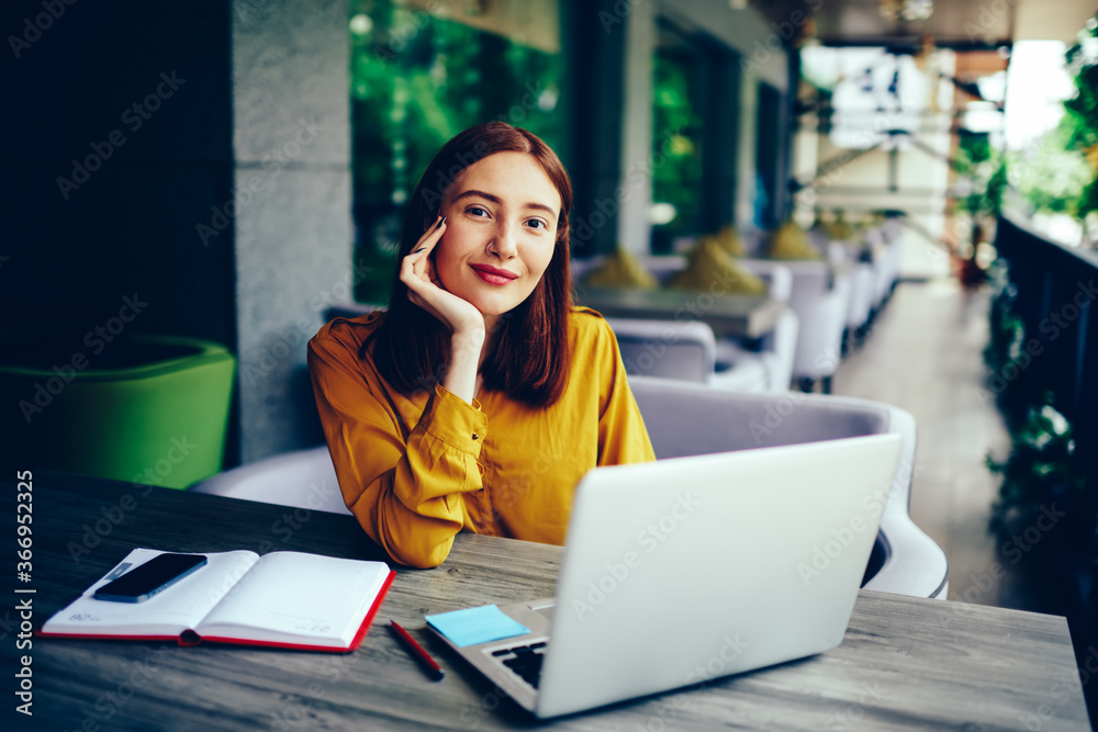 Half length portrait of positive redhead woman looking at camera during remote job on laptop computer connected to wifi.Young cute female student sitting at wooden table with gadgets and notepad Half length portrait of positive redhead woman looking at camera during remote job on laptop computer connected to wifi.Young cute female student sitting at wooden table with gadgets and notepad