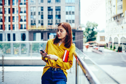 Young attractive hipster girl checking notification on smartphone while strolling at street in sunny day.Charming female blogger sharing news with followers in social network via modern cellphone