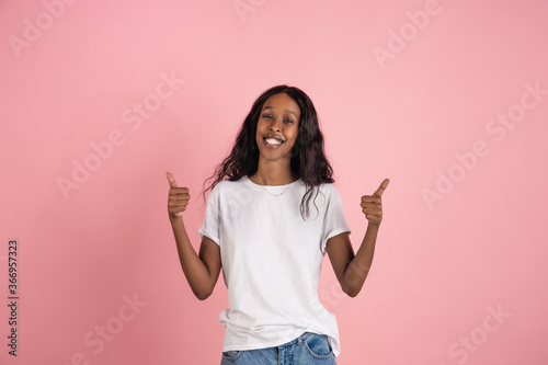 Thumbs up. Cheerful african-american young woman isolated on pink background, emotional and expressive. Concept of human emotions, facial expression, sales, ad. Beautiful model with long curly hair.