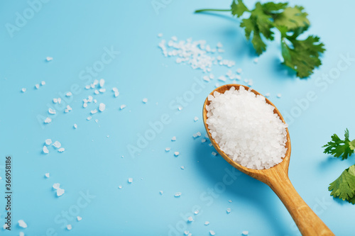 Large white sea salt in a wooden spoon on a blue background, with salt and herbs scattered around the edges.