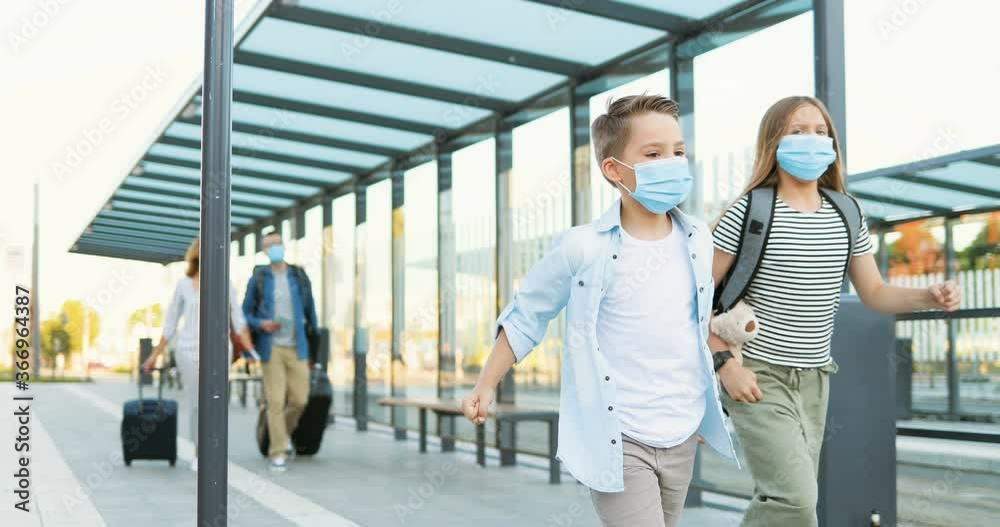 Cute small Caucasian happy kids, brother and sister in medical masks running at train station. Parents walking on blurred background and carrying suitcases on wheels. Trip concept in pandemic. Cute small Caucasian happy kids, brother and sister in medical masks running at train station. Parents walking on blurred background and carrying suitcases on wheels. Trip concept in pandemic.