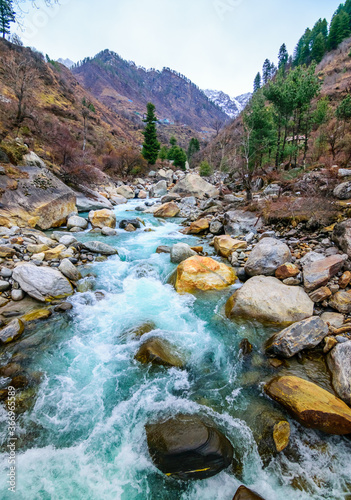 A flowing river with rocks on its way and by the sides. Mountain and pine trees visible in the background