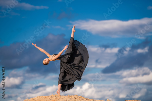 Tableau sur toile young ballerina in a black long dress hovers above the ground like a bird, resisting a gust of wind against a background of blue sky and clouds