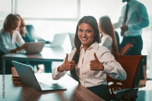 Businesswoman shows thumbs up in office. Alright gesture from young smiling girl. Group of businesspeople work on blurred background. Achievement in business concept. Tinted image.
