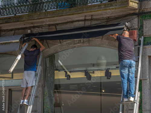 Two Men On Ladders, Hanging An Awning