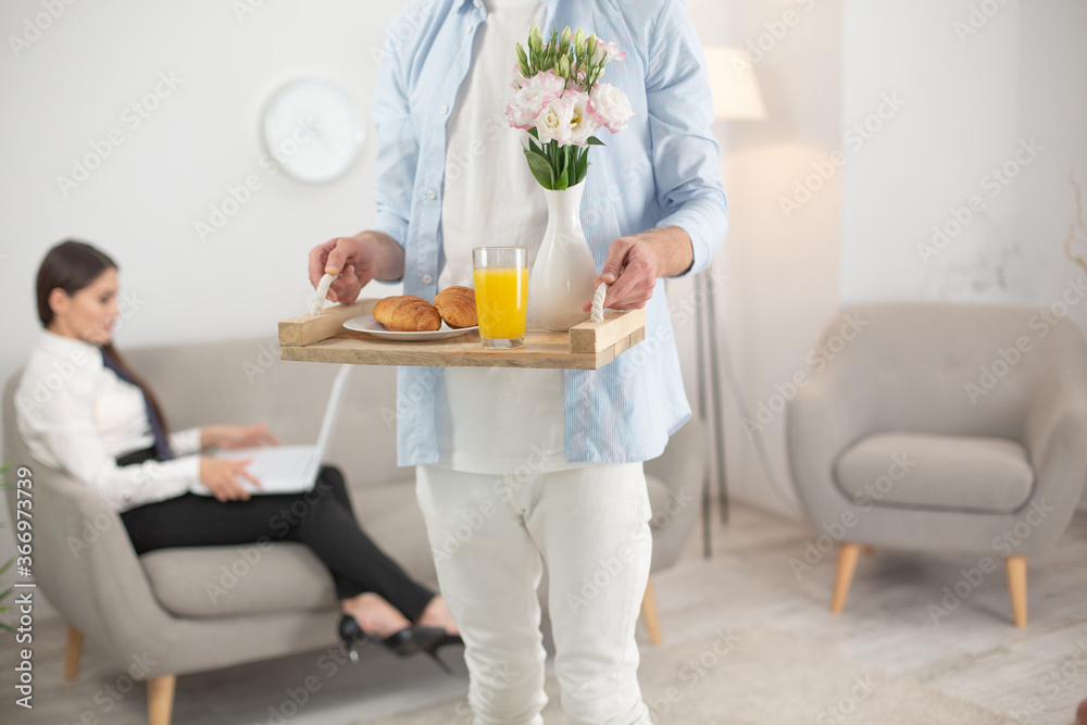 Man bringing his wife food. Cropped picture of man holding food tray. Cropped shot of male wearing white clothes holding up tray with breakfast food on it. Woman sits at sofa on blurred background. Man bringing his wife food. Cropped picture of man holding food tray. Cropped shot of male wearing white clothes holding up tray with breakfast food on it. Woman sits at sofa on blurred background.