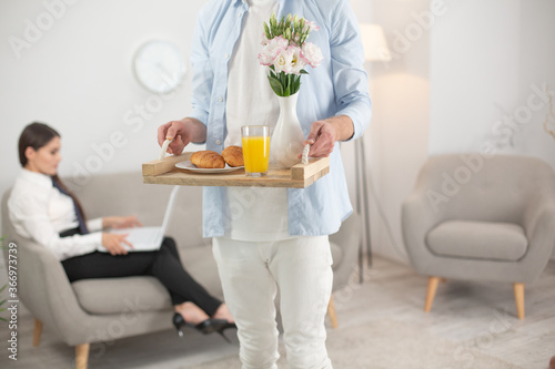 Man bringing his wife food. Cropped picture of man holding food tray. Cropped shot of male wearing white clothes holding up tray with breakfast food on it. Woman sits at sofa on blurred background.