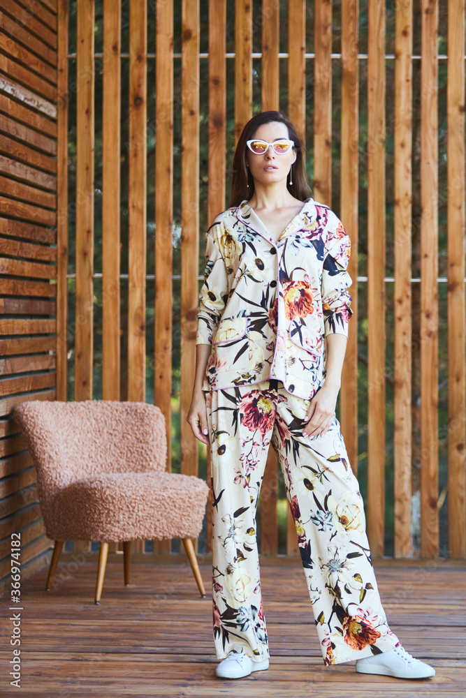 Young stylish sexy woman in pajamas standing on terrace in tropical hotel, palm trees background, long black hair, sunglasses, earrings, sunglasses.