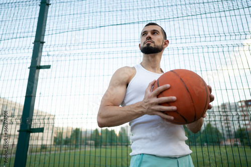 Man stands near a fence on the street, holding a basketball in his hand