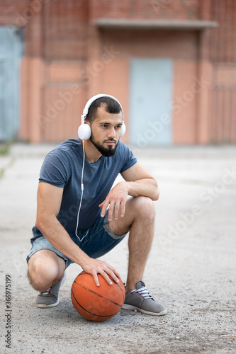 Man with headphones on the street is preparing to play basketball, sitting holding a basketball in his hand