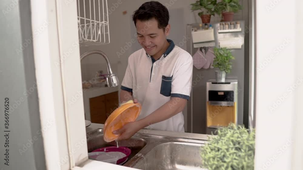 Happy young asian man in white shirt standing and washing dishes on the kitchen Happy young asian man in white shirt standing and washing dishes on the kitchen