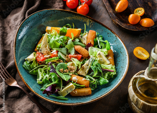 Fresh summer salad with deep-fried cheese sticks, blue cheese, tomatoes, cucumbers, granola, greens and orange in bowl on wooden background. Healthy food, clean eating, Buddha bowl salad, top view