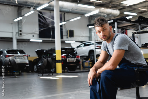 tired technician in overalls holding cap and looking at camera while sitting near cars in workshop
