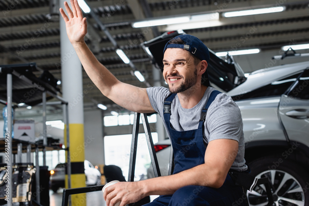 happy mechanic in cap waving hand and holding paper cup in workshop