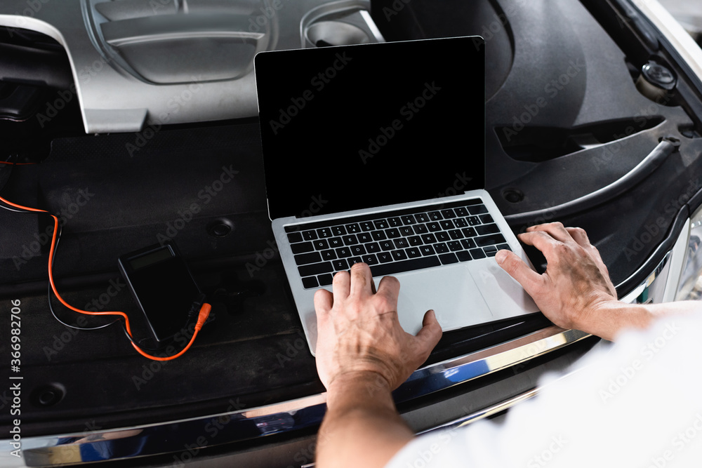 Cropped view of auto mechanic using laptop on car near wires and multimeter at service station Cropped view of auto mechanic using laptop on car near wires and multimeter at service station