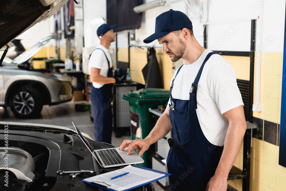 Selective focus of auto mechanic using laptop on car near clipboard at service station