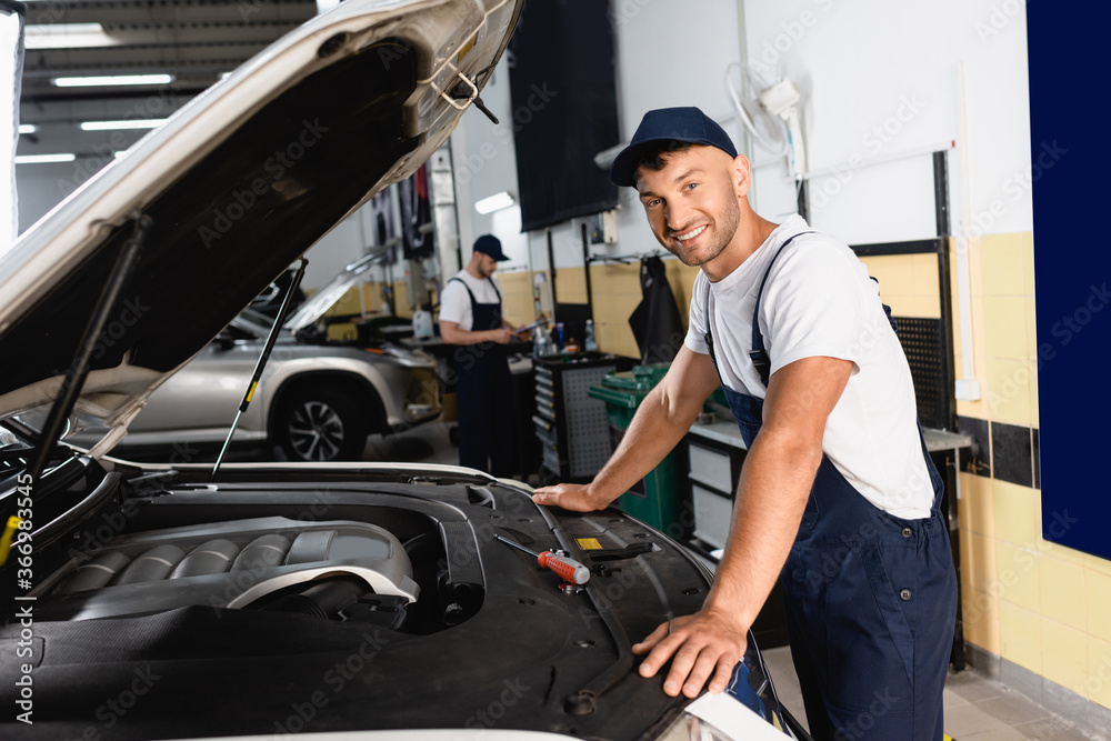 selective focus of happy mechanic in cap smiling near car and coworker in workshop selective focus of happy mechanic in cap smiling near car and coworker in workshop