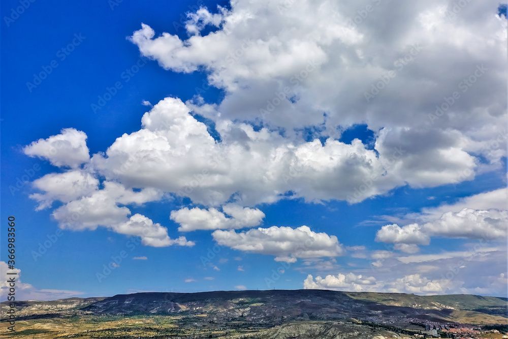 Beautiful white cumulus clouds in the blue sky over Cappadocia. Below is a valley, hills, village. Summer sunny day. Beautiful white cumulus clouds in the blue sky over Cappadocia. Below is a valley, hills, village. Summer sunny day.
