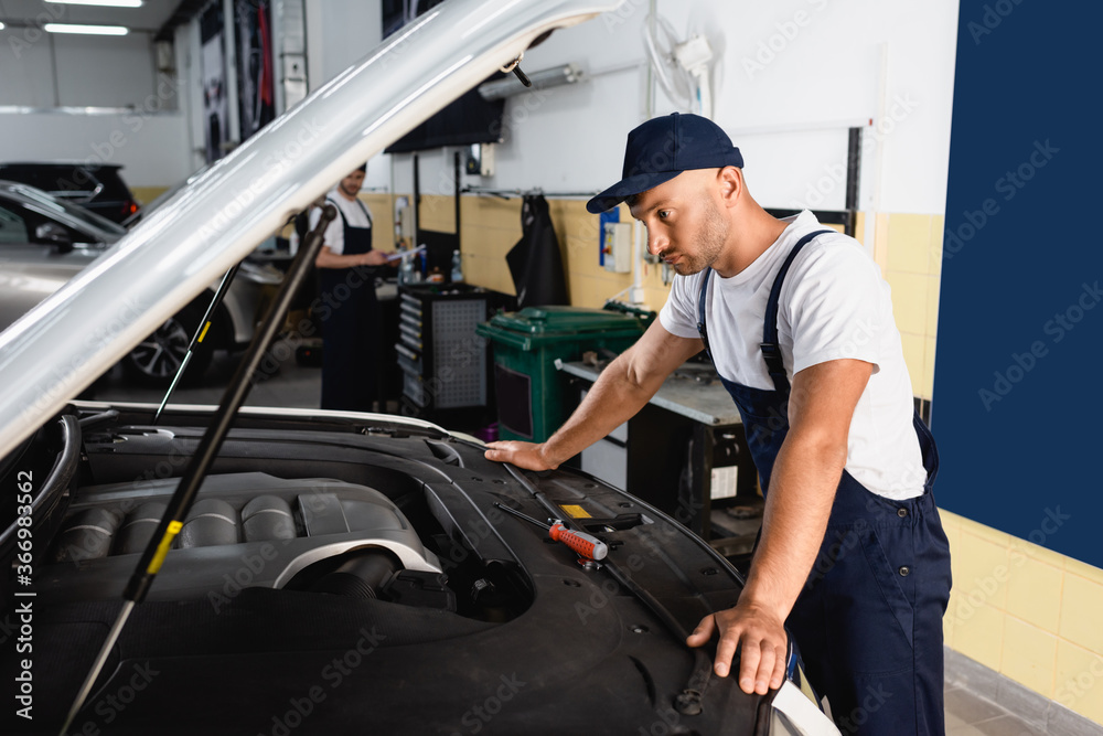 selective focus of mechanic in cap looking at car near coworker in workshop