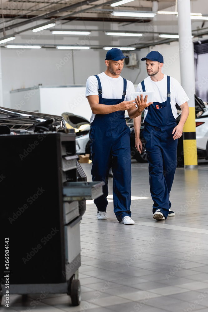 selective focus of handsome mechanics in uniform using digital tablet and walking in workshop selective focus of handsome mechanics in uniform using digital tablet and walking in workshop