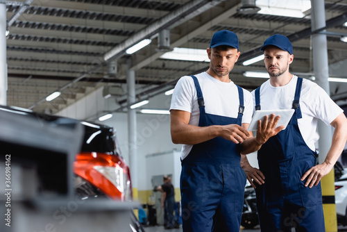 selective focus of handsome mechanics in uniform using digital tablet and looking at car in workshop