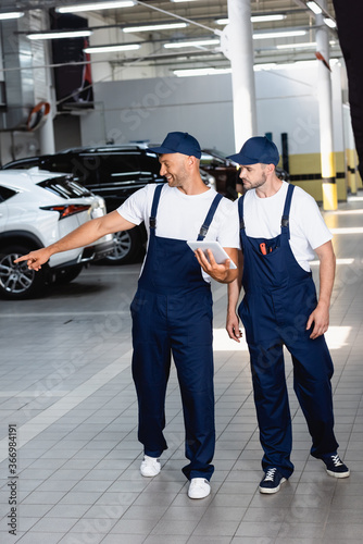 happy mechanic in uniform holding digital tablet and pointing with finger at car near coworker in workshop