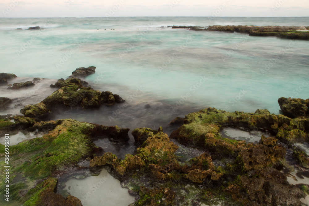 Long exposure shot of the beach at sunset. View of the shore mossy rocks,  turquoise ocean water and blurred sea waves creating a smoky effect with a beautiful dusk light.