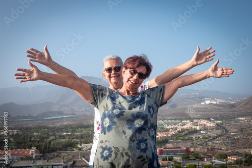 Mature couple of women standing on hill with mountain and city on background looking at camera with arms raised - active seniors and freedom concept