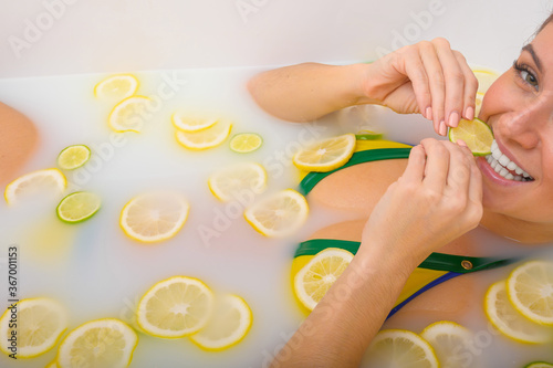 Beautiful young red-haired woman takes a bath with milk and citrus fruits. Portrait of Caucasian smiling girl eating lemon wedge in skin whitening bath. Taking care of the body.
