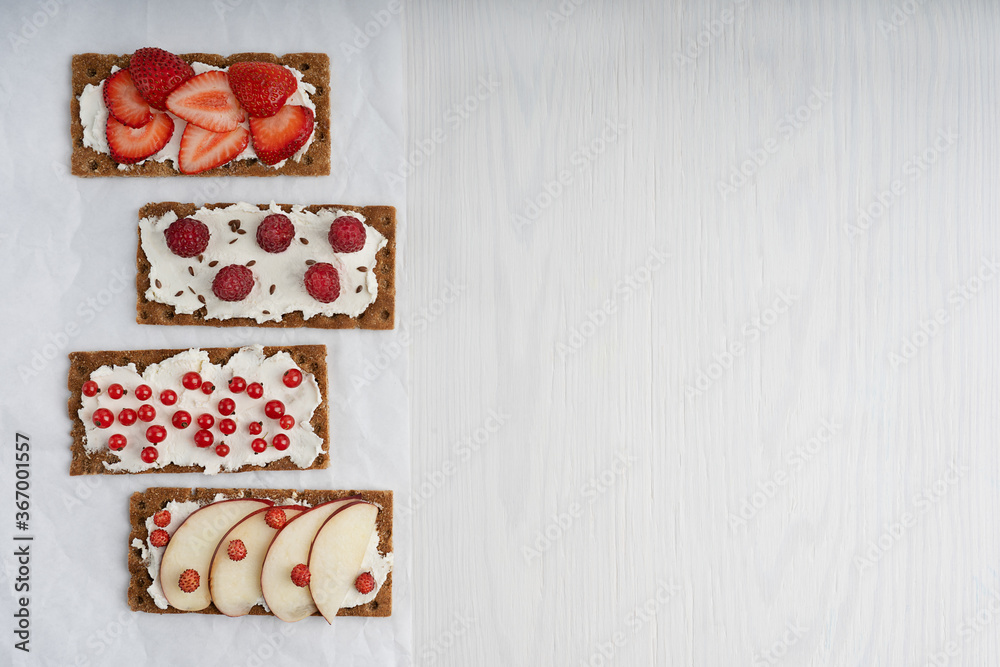 Top view of four pieces of vegetarian crisp bread with cream cheese and assortment of red berries such as strawberry, raspberry and red currant laying on white wooden table. Image with copy space Top view of four pieces of vegetarian crisp bread with cream cheese and assortment of red berries such as strawberry, raspberry and red currant laying on white wooden table. Image with copy space