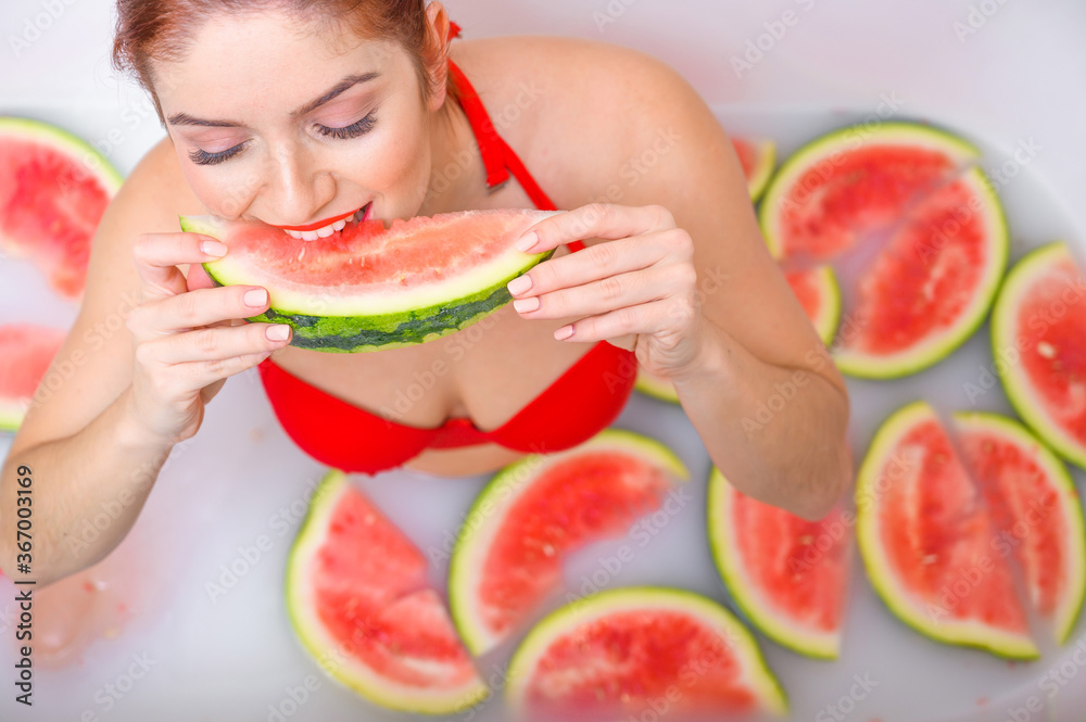 Portrait of a woman in a red swimsuit eating a watermelon and smirking. Redhead girl with red lipstick takes a bath with milk and fruit to rejuvenate her skin. Body care. Portrait of a woman in a red swimsuit eating a watermelon and smirking. Redhead girl with red lipstick takes a bath with milk and fruit to rejuvenate her skin. Body care.