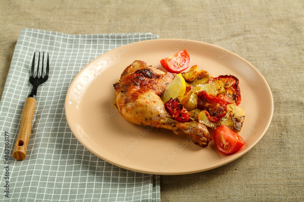 Baked chicken leg with potatoes and tomato on a beige plate on a tablecloth next to a fork and napkin. Baked chicken leg with potatoes and tomato on a beige plate on a tablecloth next to a fork and napkin.