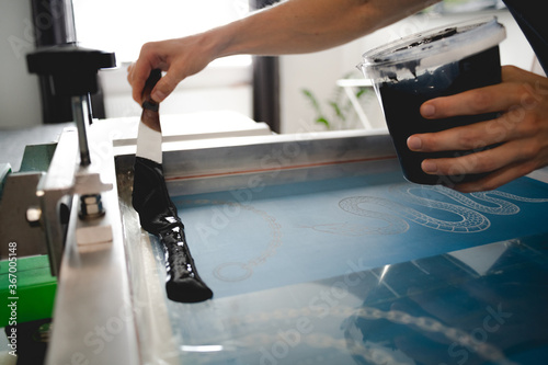 Fotografia Cropped view of artisan applying plastisol ink with spatula on stencil on screen