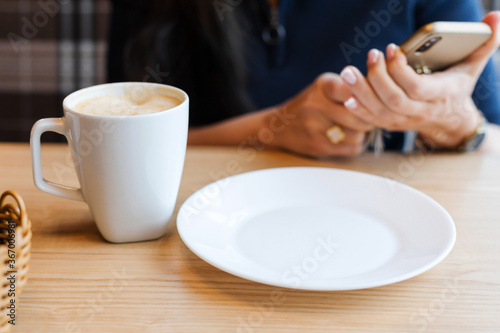 A white coffee Cup and an empty plate on a wooden table against the background girl's hands holding a smartphone while waiting for breakfast in the morning.