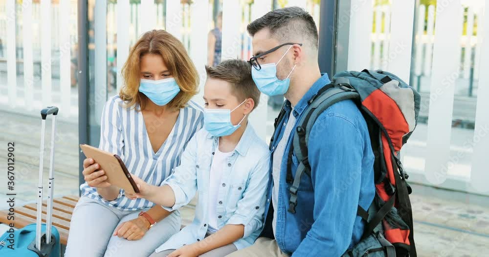Caucasian mother and father sitting on bench at bus stop with small son and using tablet device. Parents with kid in medical masks waiting for transpost with suitcases and looking for route on gadget.