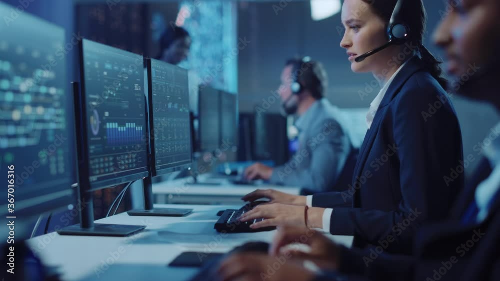 Happy Beautiful Technical Customer Support Specialist Having a Headset Call while Working on a Computer in a Dark Monitoring and Control Room Filled with Colleagues and Display Screens.