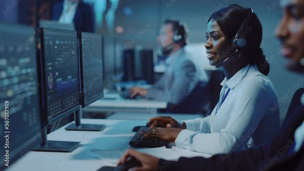Pretty Black Technical Customer Support Specialist is Talking on a Headset while Working on a Computer in aCall Center Monitoring and Control Room Filled with Colleagues and Display Screens.