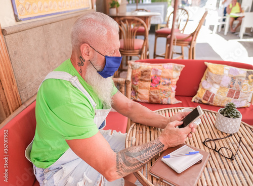 Trendy hipster with smartphone sitting in a bar - Senior man enjoy technology and wearing protective face mask - Coronvirus lifestyle photo