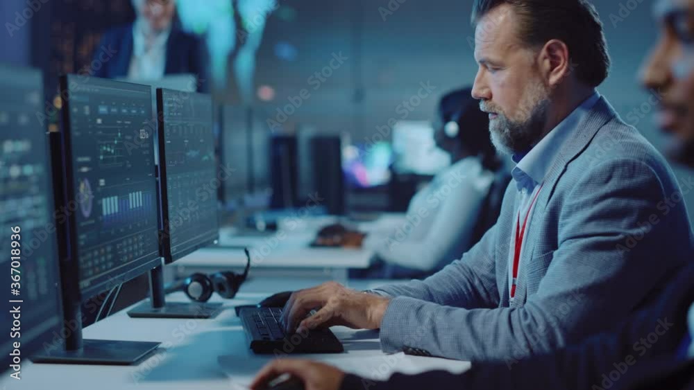 Portrait of Serious Professional Technical Controller Sitting at His Desk with Multiple Computer Displays Before Him. In the Background His Colleagues Working in System Control and Monitoring Center.