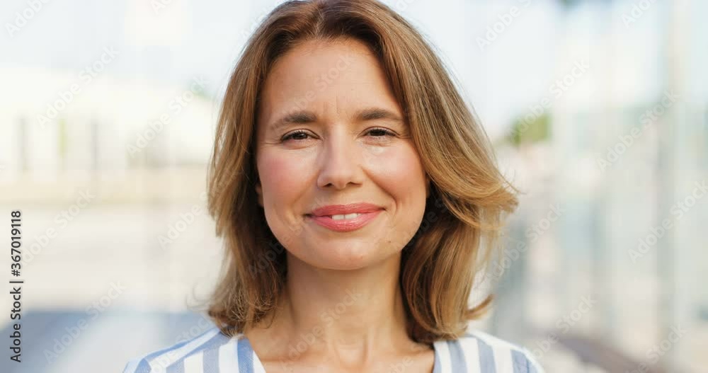 Portrait of beautiful Caucasian woman with fair hair looking at side, turning face to camera and smiling outdoors on sunny summer day. Happy female at street. Close up.
