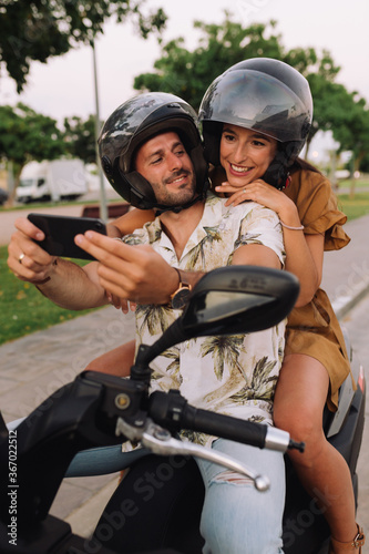 Cheerful Stylish Couple Satting On Modern Motorbike Outdoors and making a selfie with a smartphone
