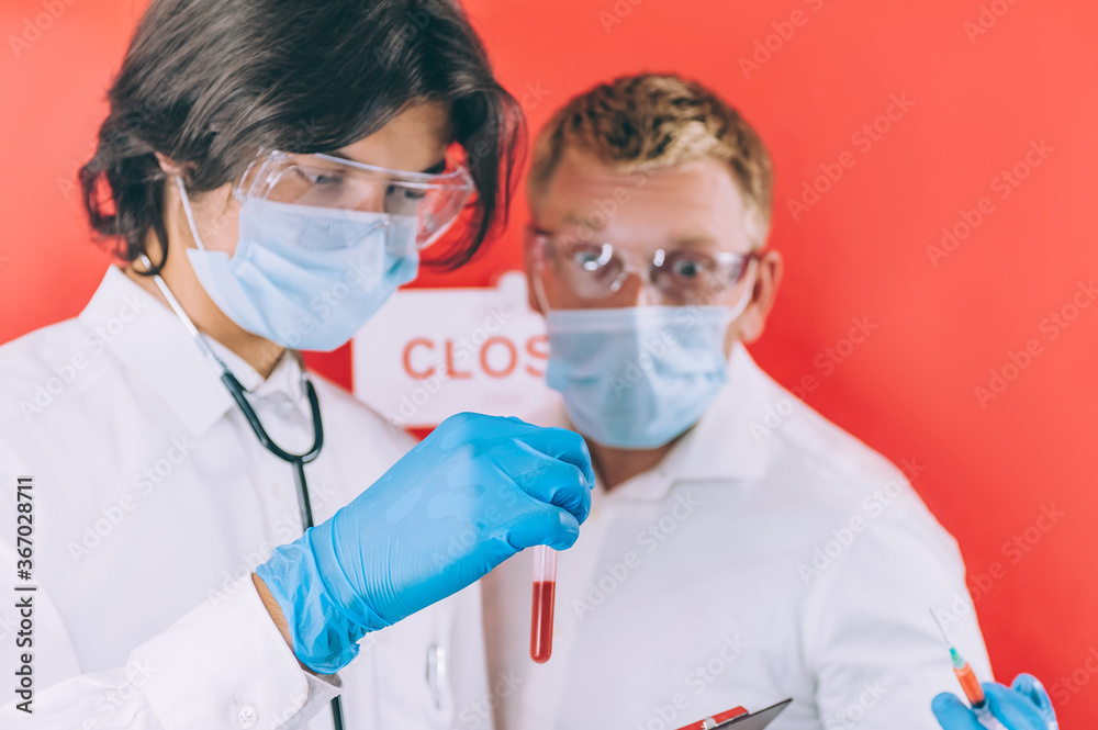 Two doctors with blue gloves, face masks and white coats check a blood test in a test tube against a red background.