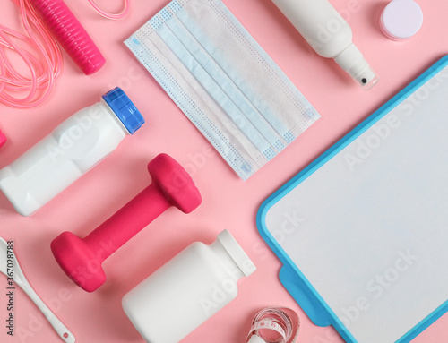 Sporting goods on a pink background.Sports items: dumbbell, measuring tape, protein drink, skipping rope, disinfectant, mask and writing board lie on a pink background, top view close up.