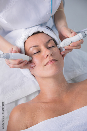 Young woman lying on cosmetologist's table during rejuvenation procedure. Cosmetologist take care about neck and face skin youthfull and wellness. Hardware face cleaning procedure. photo