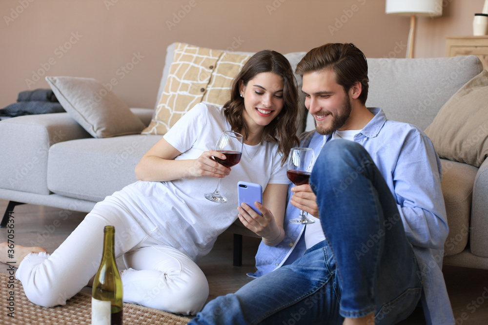 Happy couple sitting, relaxing on floor in living room, drinking red wine. Smiling young husband and wife rest at home enjoy romantic date on family weekend together