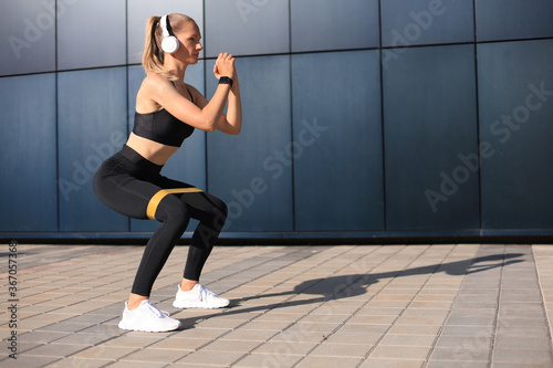Sporty woman doing squats with fitness gum expander outdoors.