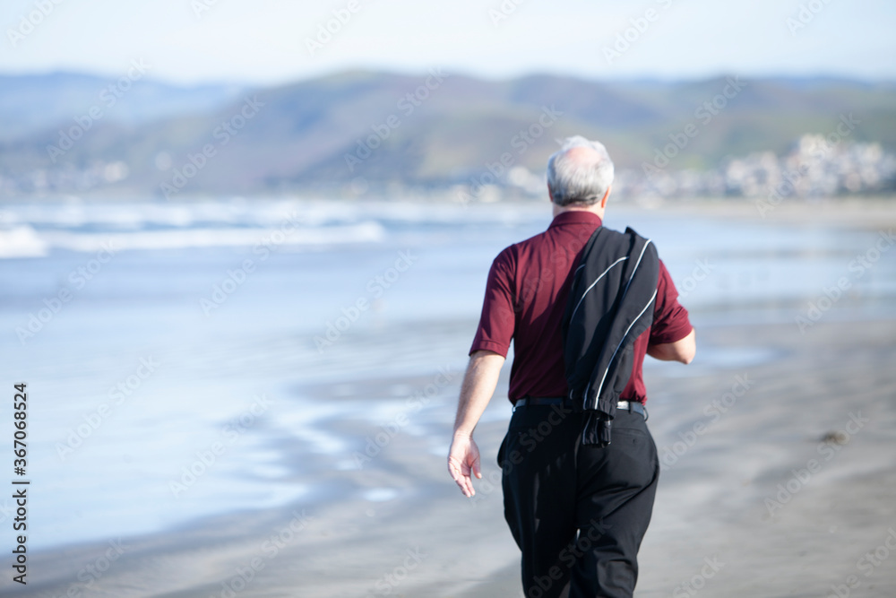 Older gentleman walking on beach with back to camera and sweater over shoulder Older gentleman walking on beach with back to camera and sweater over shoulder