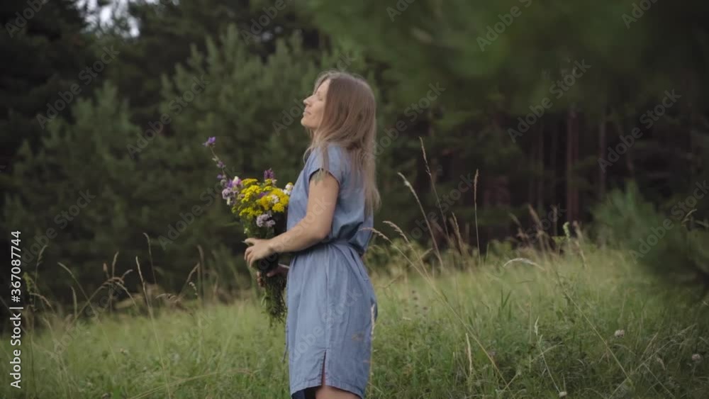 A blonde woman in a denim dress with wild flowers walks through a summer field with high green grass on a rural hill. A blonde woman in a denim dress with wild flowers walks through a summer field with high green grass on a rural hill.